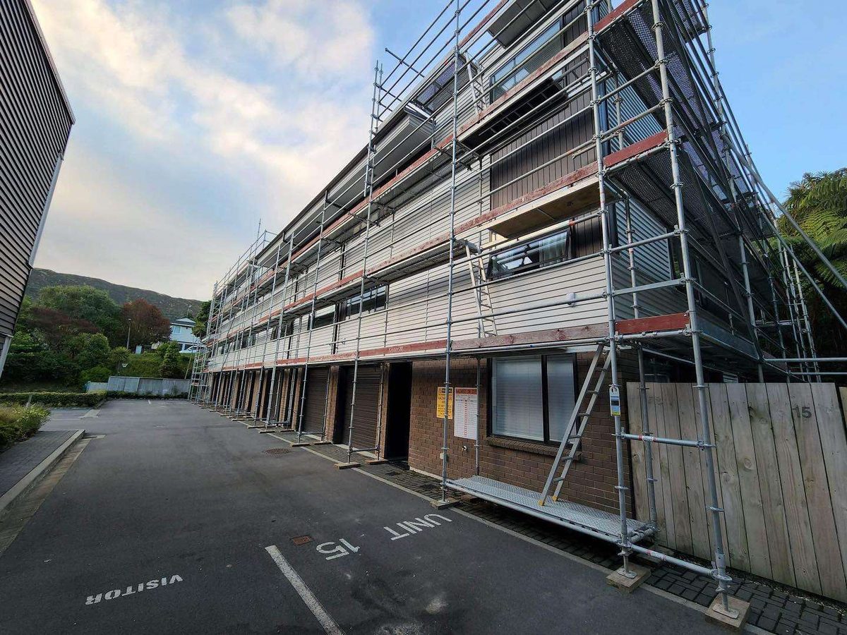 A three-storey block of units with weatherboard upper floors, covered in scaffolding along its whole length beside a car park