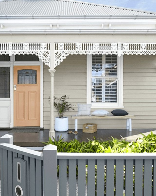 The front of a villa with pale weatherboards, a white verandah with decorative fretwork, a timber front door and a grey picket fence