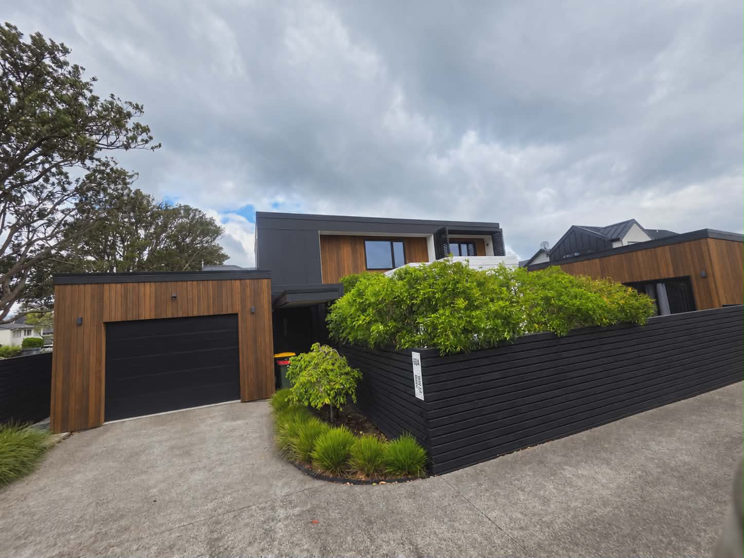 A garage clad in vertical stained cedar boards with a black door, beside a black horizontal-board fence and planting on a concrete driveway, a two-storey house behind