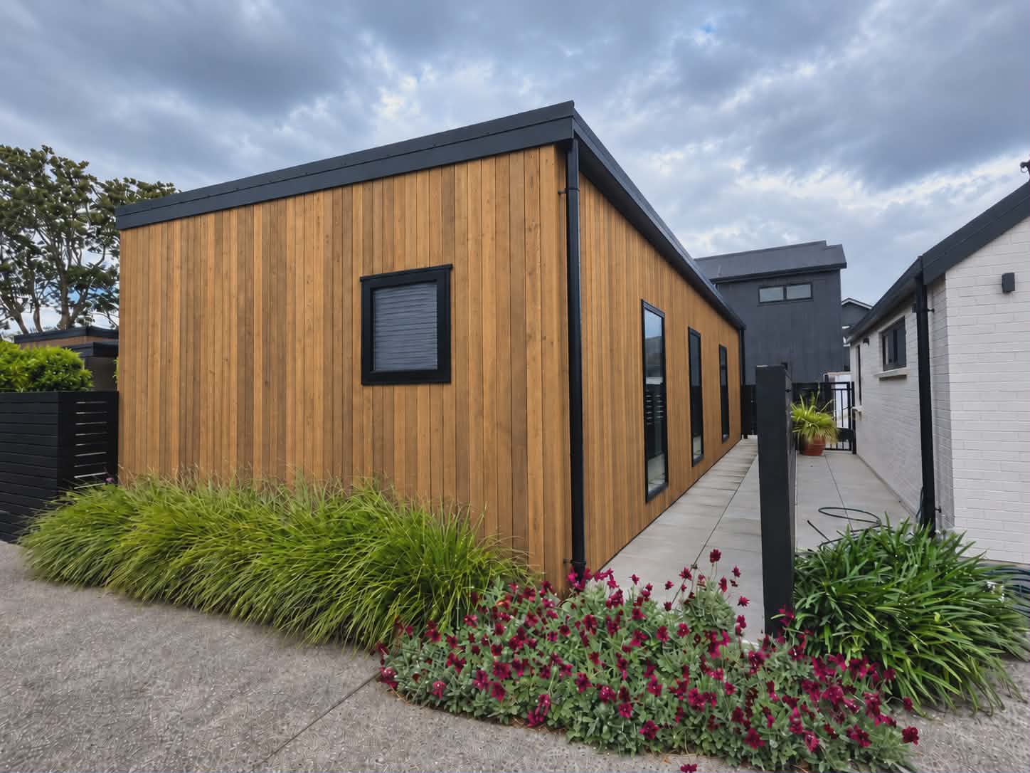 The side of a building clad in vertical stained cedar boards with black-framed windows, behind a garden of grasses and red flowers beside a concrete path