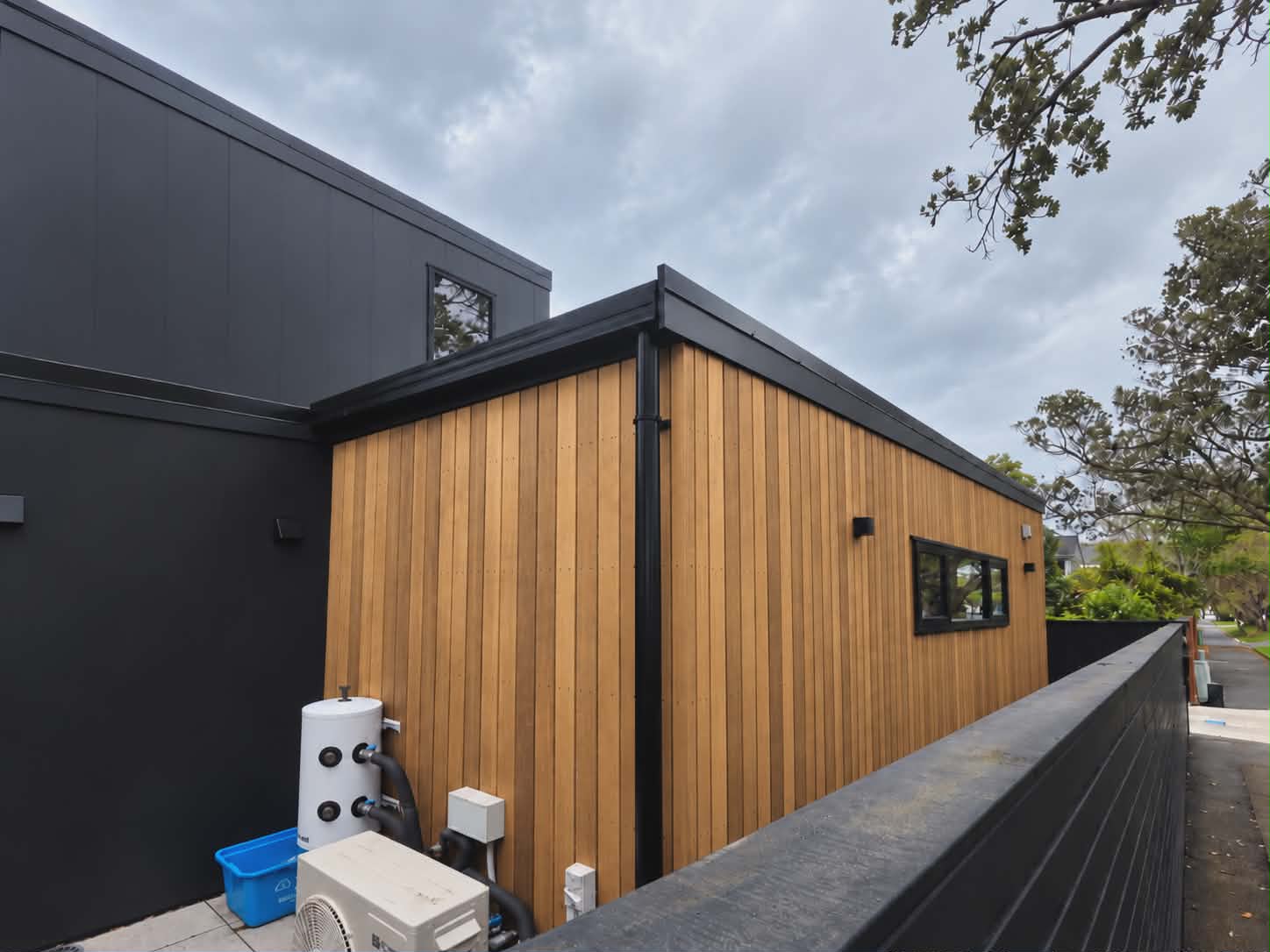 A single-storey wing clad in vertical stained cedar boards with a black-framed window, joined to a dark two-storey house, with a heat pump unit and hot water cylinder beside a black fence