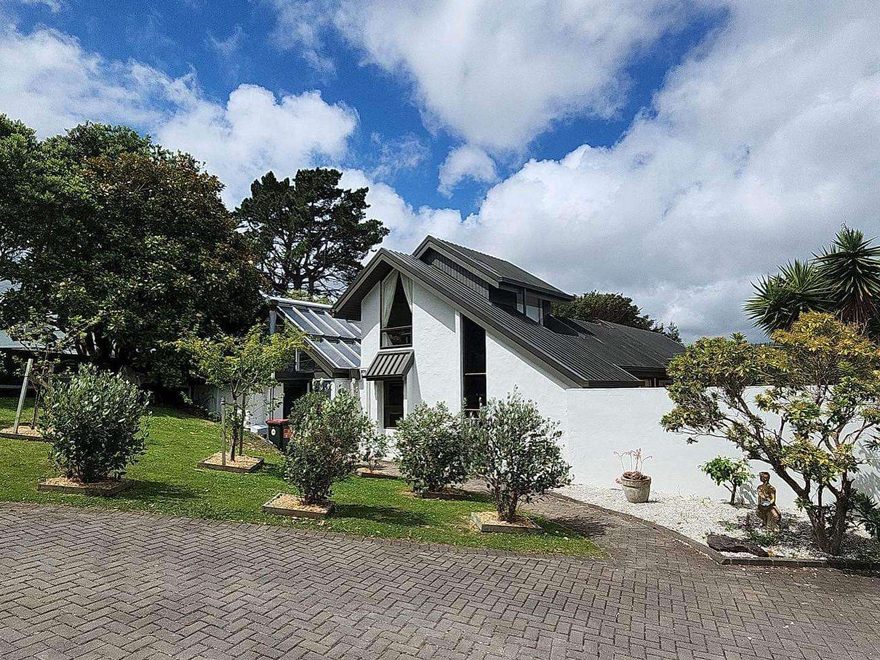 A white house with steep dark grey roofs behind a paved courtyard planted with olive trees